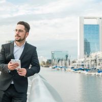 portrait-young-businessman-standing-near-harbor-holding-mobile-phone-hand-looking-away (1)